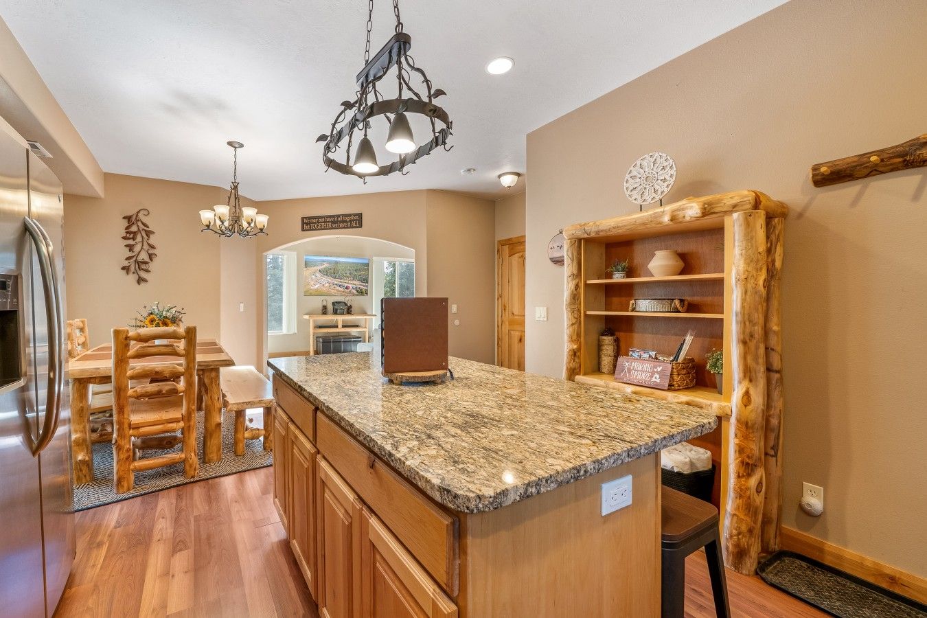 A kitchen with a granite countertop, wooden cabinets, a refrigerator, a dining area with a wooden table and chairs, and a decorative wall piece.