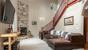 A spacious living room with a stone fireplace, a wooden coffee table, a large sectional sofa, and a wooden staircase. - PET-FRIENDLY FOREST RETREAT CABIN | HOT TUB, FIRE-PIT, & A/C (Duck Creek Village)