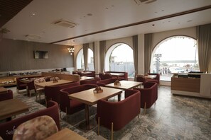 Un restaurant modern amb taules altes i cadires tapissades de color vermell, grans finestres amb cortines i una zona de cuina oberta.
