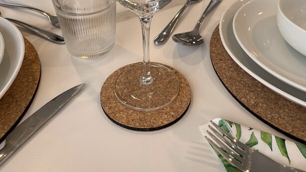 Una mesa de comedor preparada con cristalería transparente, platos y cubiertos, adornada con posavasos de corcho y un mantel individual estampado.