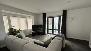 Un salón con un sofá gris, un televisor de pantalla plana sobre un soporte negro, una planta en maceta y un ventanal corredero que da a un área exterior.