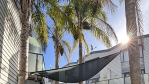 Una mesa de madera con sillas, una parrilla y un área sombreada en un patio trasero con palmeras y una casa al fondo.