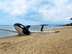 Two orca sculptures stand on the beach, facing the ocean, near a white fence and a distant city skyline.