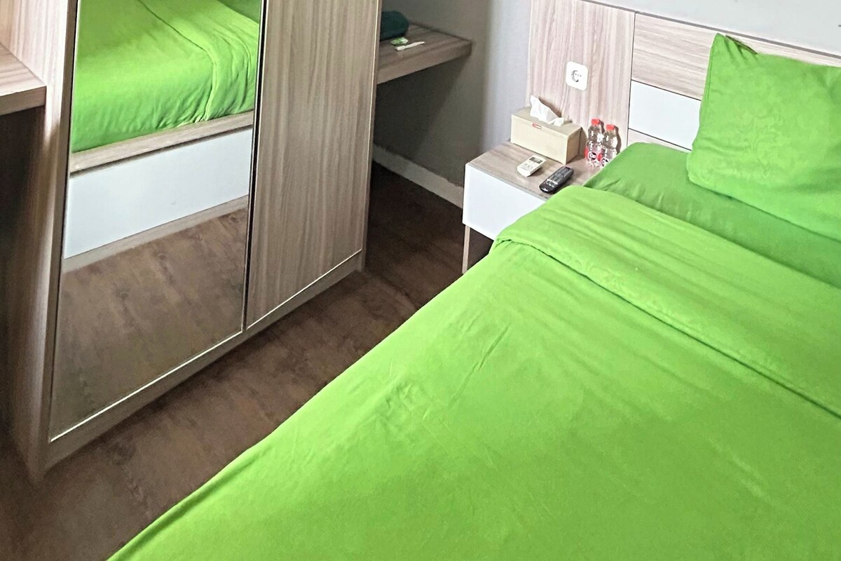 Una habitación de hotel con una cama, una mesita de noche con un control remoto, un espejo y un pequeño estante con artículos de aseo.