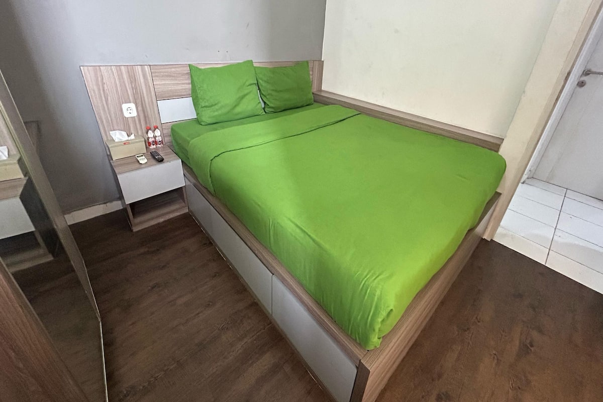 A single bed with a green bedspread in a room with wooden flooring and a white wall.