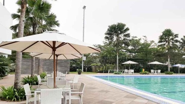 Zona junto a la piscina con sillas y sombrillas blancas, rodeada de palmeras y una pileta.