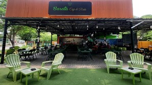 Un espacio al aire libre con sillas y mesas verdes, un porche cubierto y un letrero que dice 'Barata Cafe & Resto'.