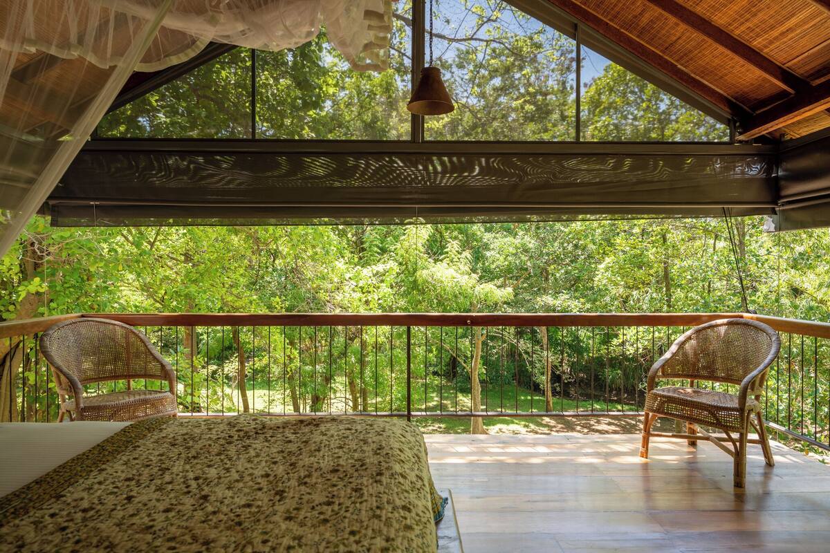 Uma varanda com cama e duas cadeiras de vime, com vista para uma floresta verdejante.