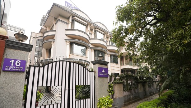 Uma entrada com portão e uma placa indicando "16 ARJUN MARG DLF PHASE-1", além de uma placa de hotel com a inscrição "STAY WELL".