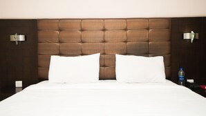Quarto de hotel com cabeceira estofada, duas almofadas brancas, uma garrafa de água e um controle remoto na mesa de cabeceira.