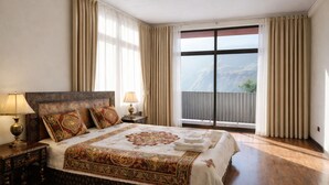 A spacious bedroom with a large bed, two bedside tables with lamps, a view of a mountain through a large window, and a balcony with curtains. - Rock Land Guest House (Lalibela)