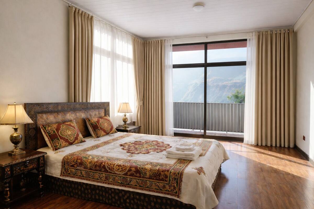 A spacious bedroom with a large bed, two bedside tables with lamps, a view of a mountain through a large window, and a balcony with curtains.