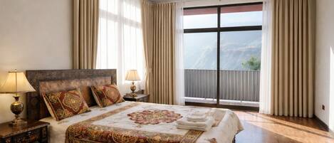 A spacious bedroom with a large bed, two bedside tables with lamps, a view of a mountain through a large window, and a balcony with curtains.