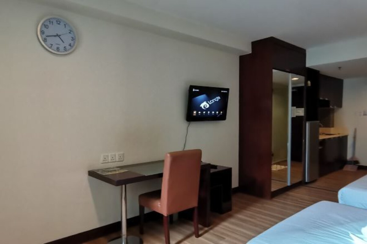 A hotel room with a desk, a chair, a bed, a TV, and a clock on the wall.