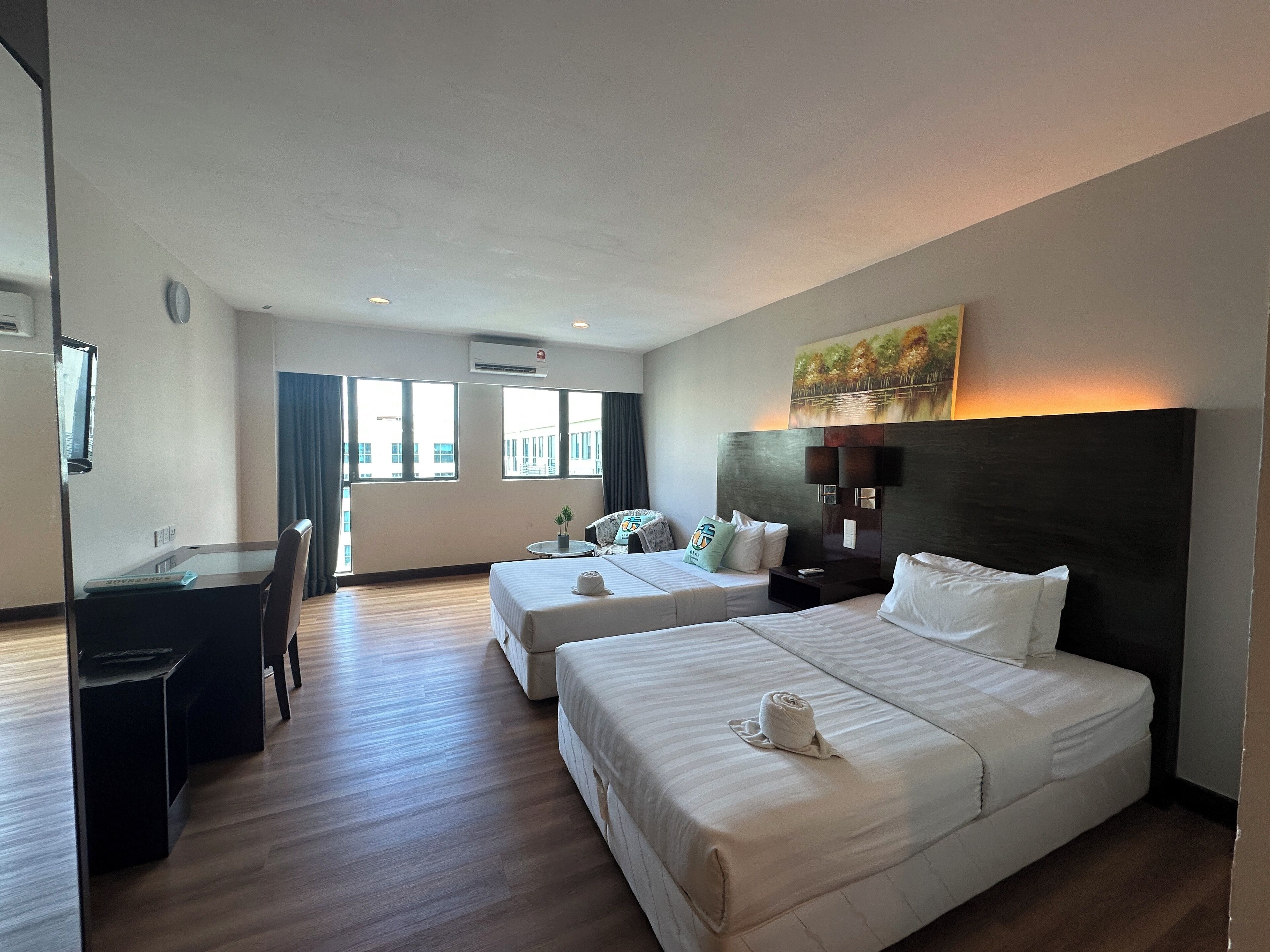 Ein Hotelzimmer mit zwei Betten, einem Schreibtisch und einem Sessel. An der Wand hängt ein Gemälde und unter der Decke befindet sich eine Klimaanlage.