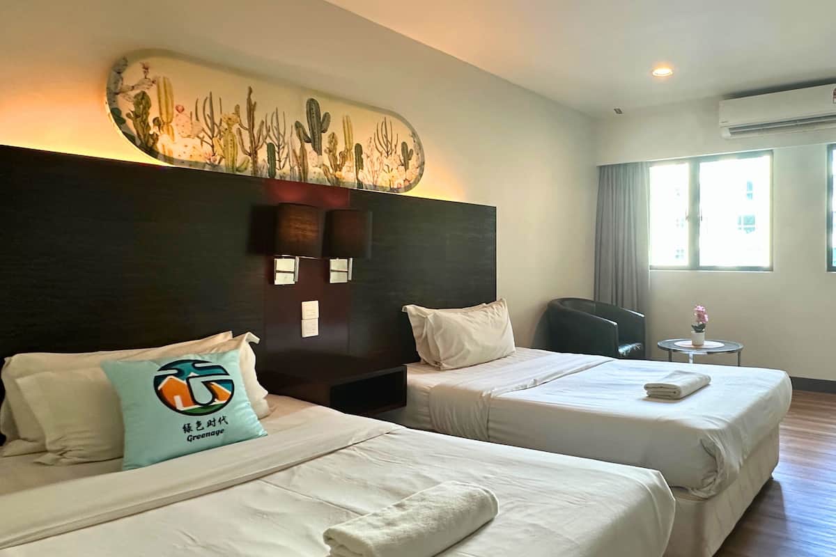 A hotel room with two beds, a dark headboard, a wall-mounted artwork, a small table with a plant, and a window with curtains.