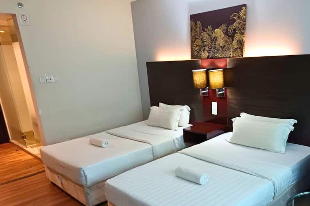 A hotel room with two single beds, a wooden floor, a dark headboard, and a wall-mounted lamp.