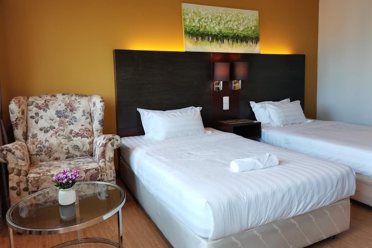 A hotel room with two beds, a floral armchair, a glass coffee table with a vase of flowers, and a painting on the wall.