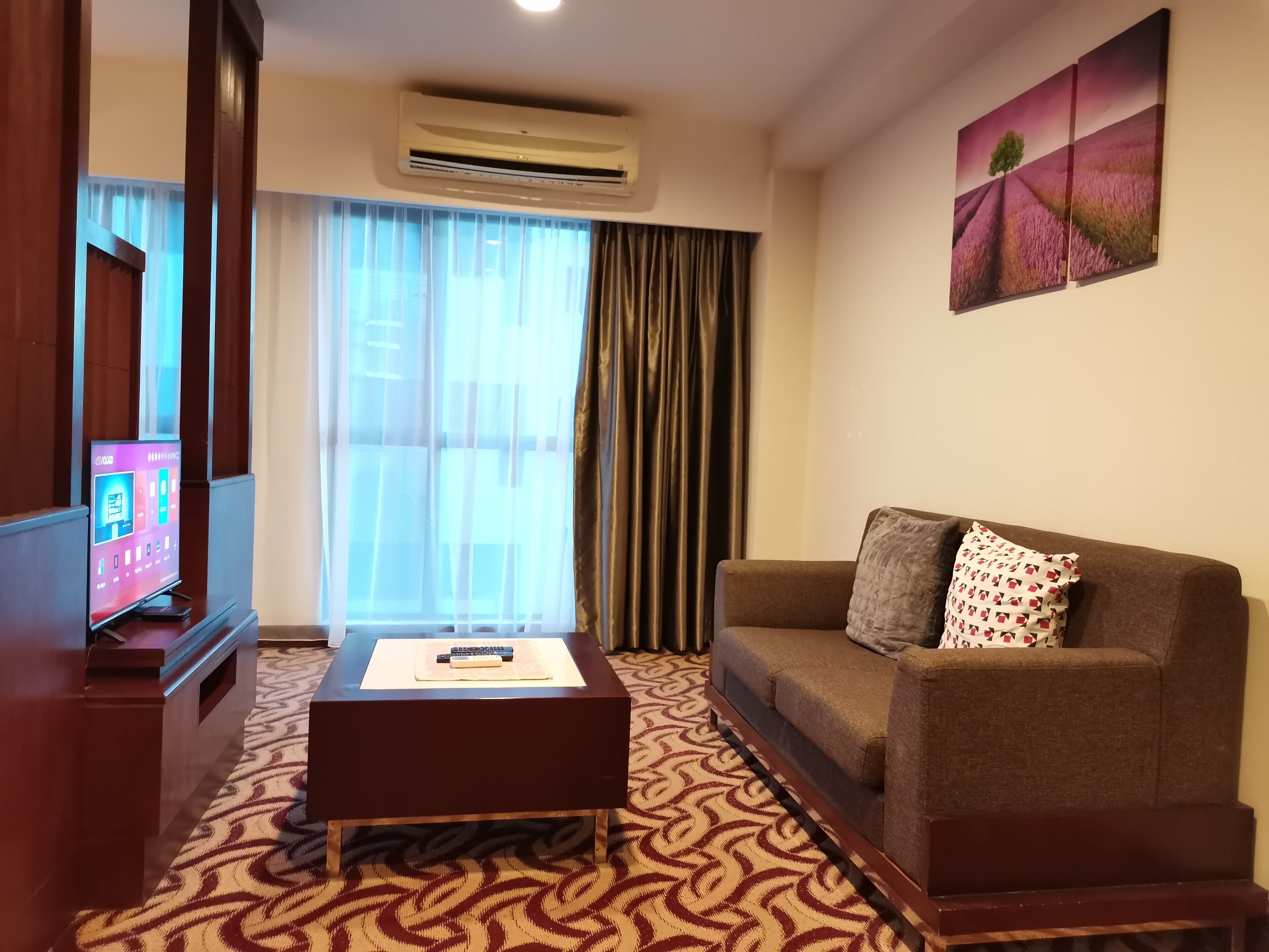 A hotel room with a sofa, a coffee table, a TV, and a large window with curtains.