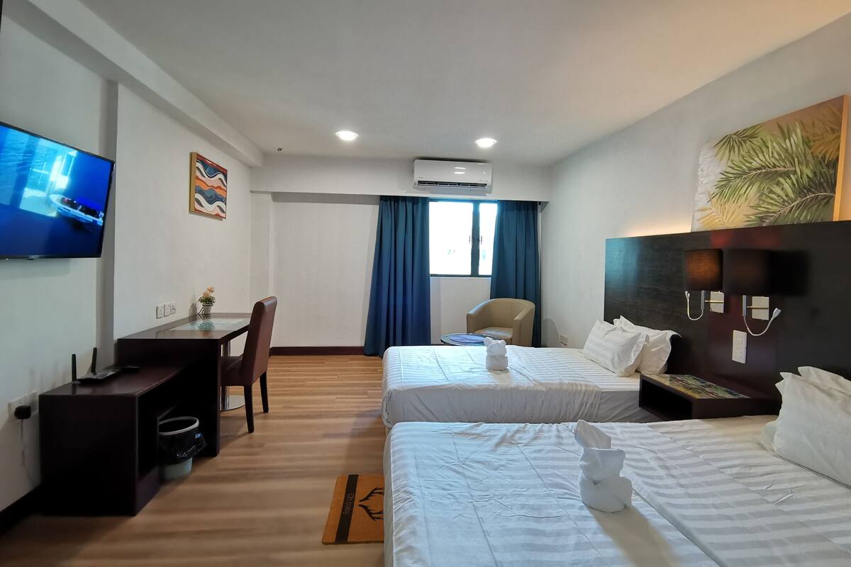 A hotel room with two beds, a desk, a chair, a TV, and an air conditioning unit.