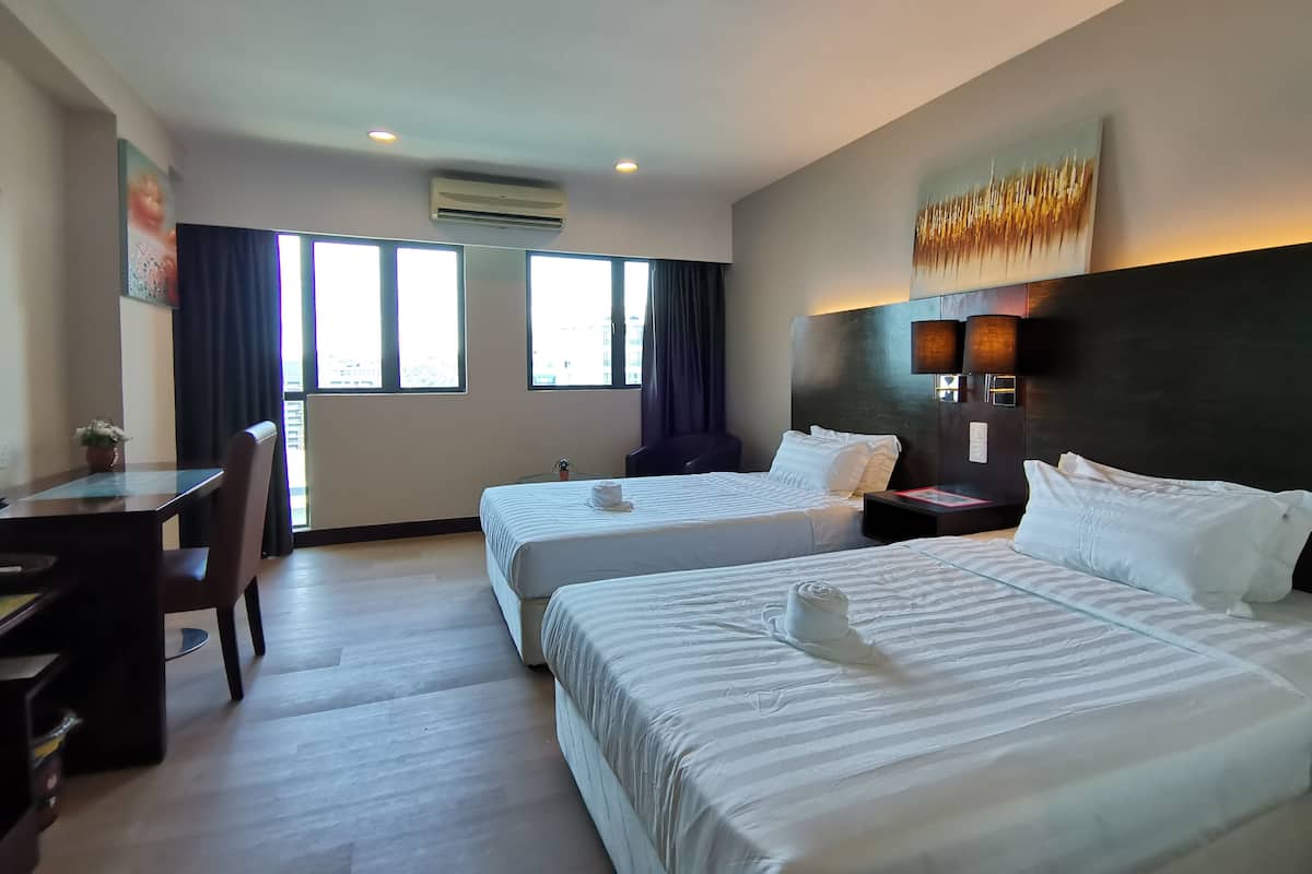 A hotel room with two beds, a desk, and a chair. There are windows with curtains, an air conditioning unit, and a painting on the wall.