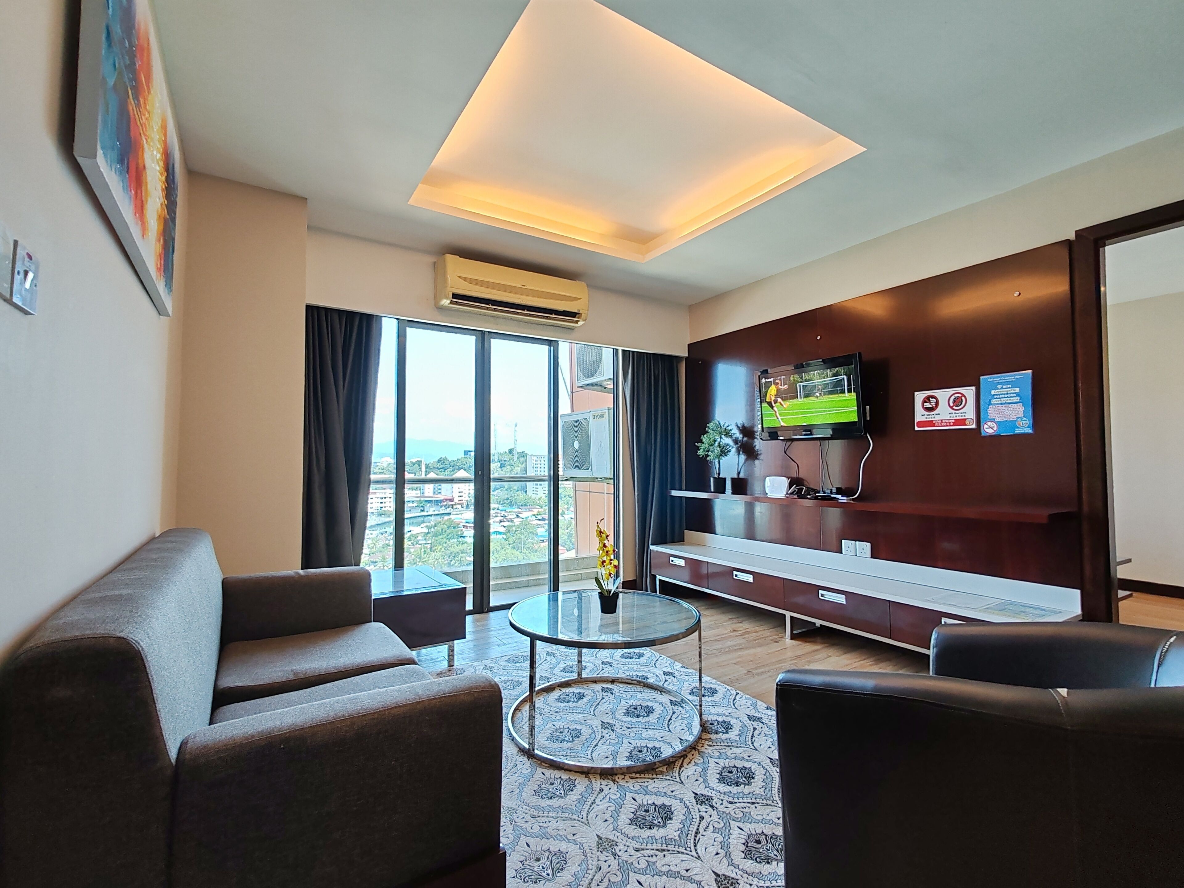 Habitació d'hotel moderna amb un sofà, una taula rodona petita, un televisor muntat a la paret i una finestra gran que ofereix vistes a la ciutat.