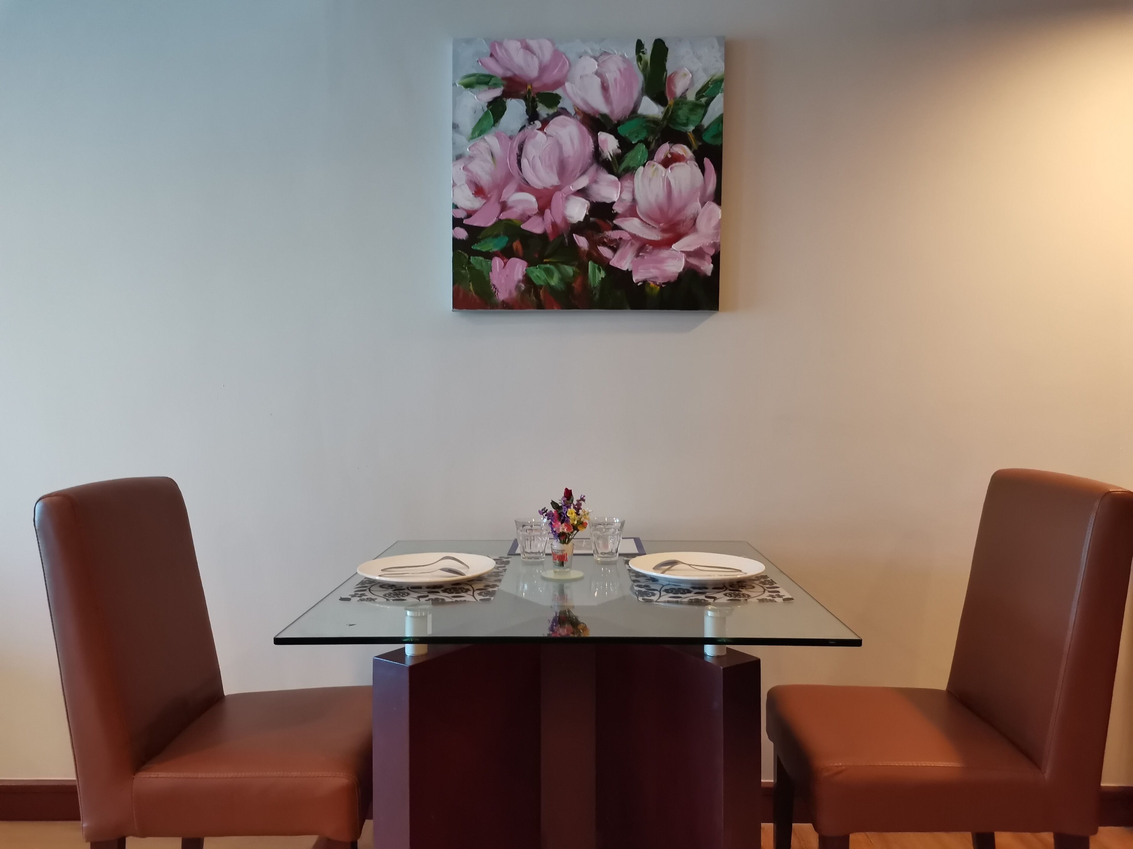 A dining table with two chairs, a glass top, and a floral painting above it.