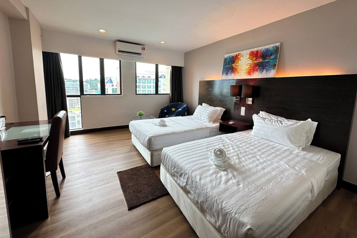 A hotel room with two beds, a desk, and a chair. There is a window with a view of buildings outside.