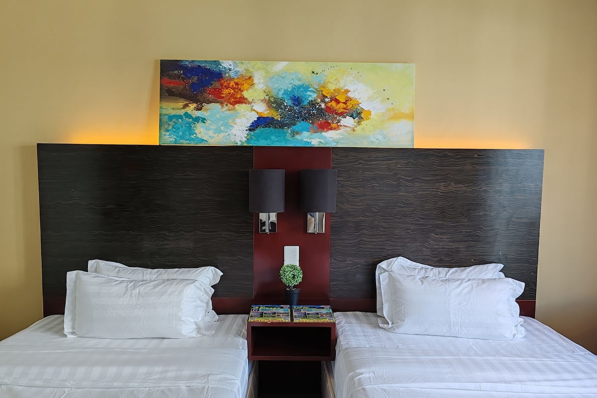 A hotel room with two beds, a large abstract painting above the beds, a dark wood headboard, and a small nightstand with a lamp and a potted plant.