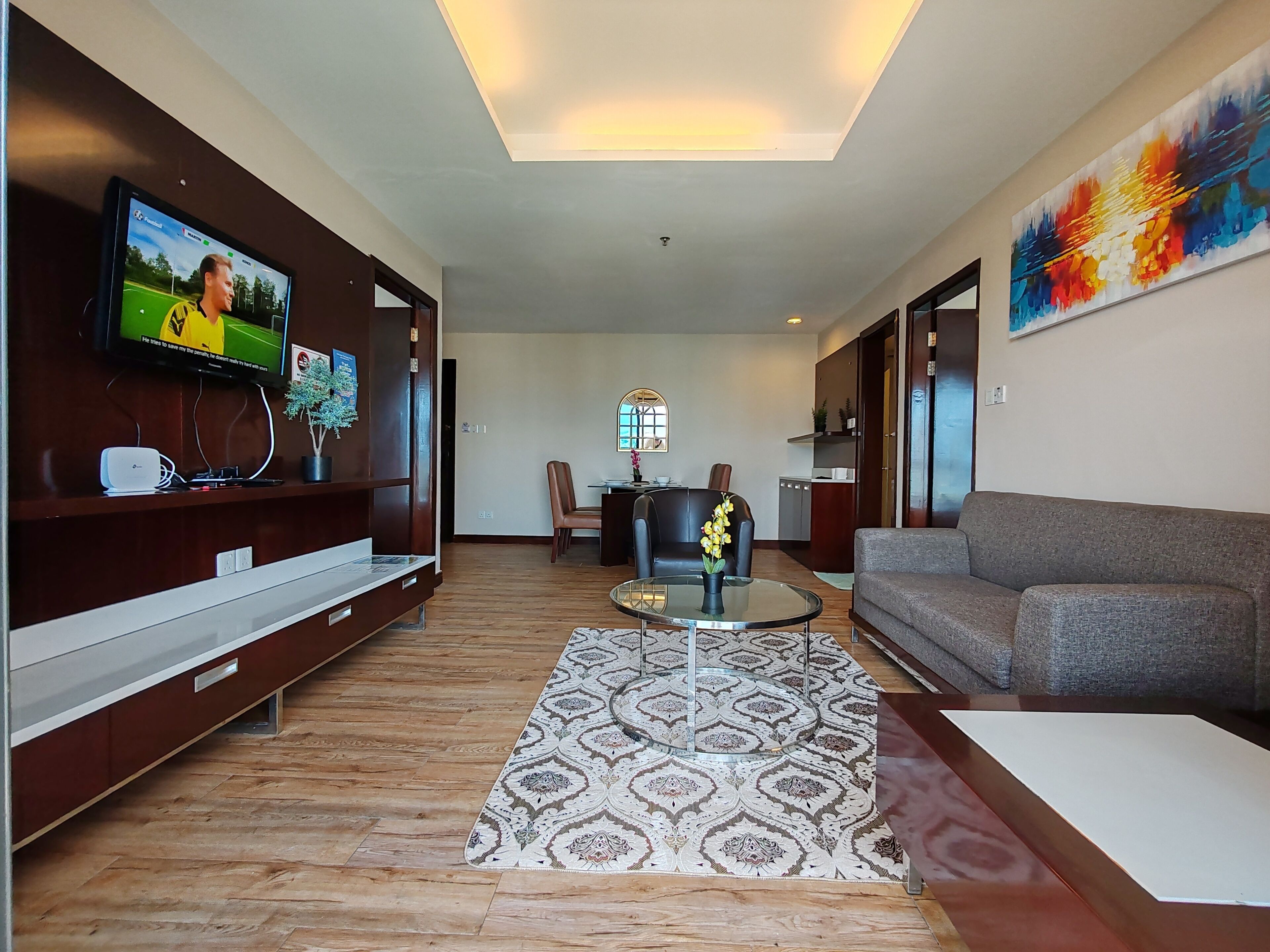 A modern living room with a flat-screen TV, a grey sofa, a patterned rug, and a dining area with a table and chairs.