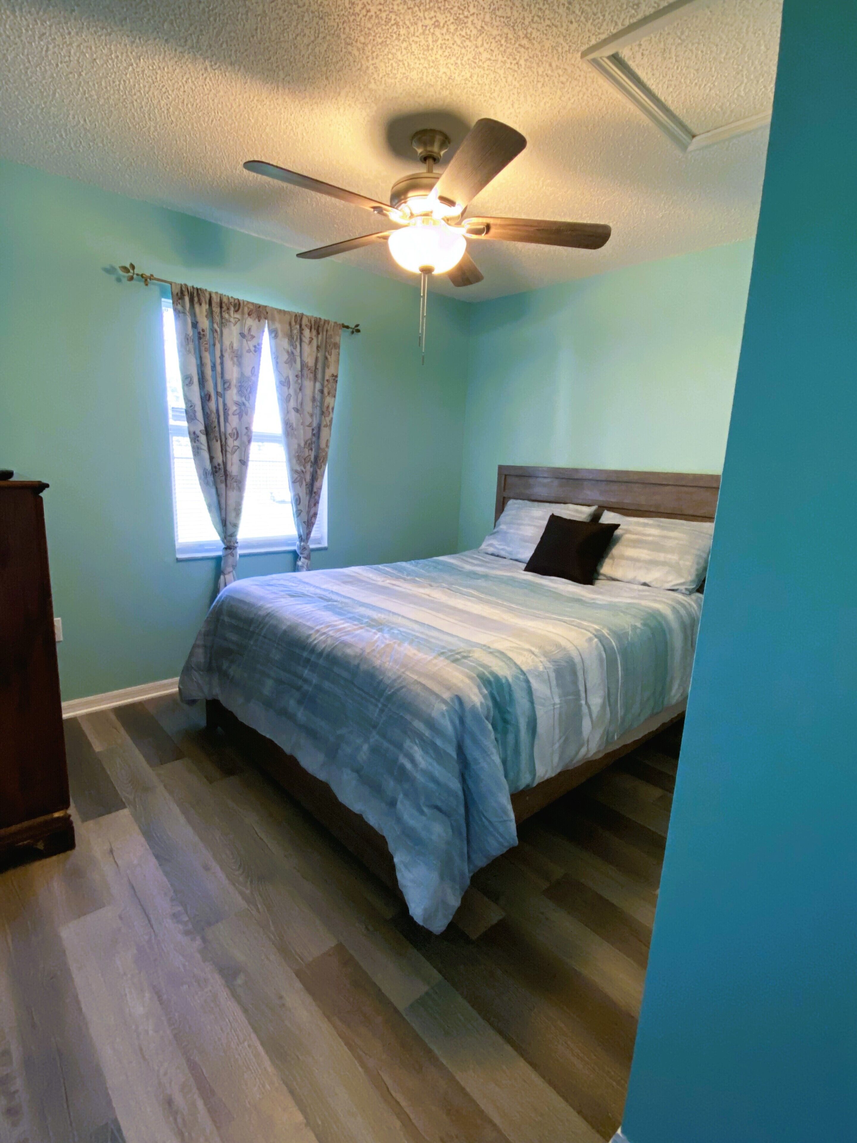 Une chambre à coucher avec un lit, un ventilateur de plafond, une fenêtre avec des rideaux et une commode en bois.