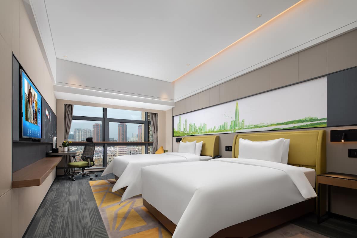 A modern hotel room with two beds, a desk, a TV, and a cityscape view.