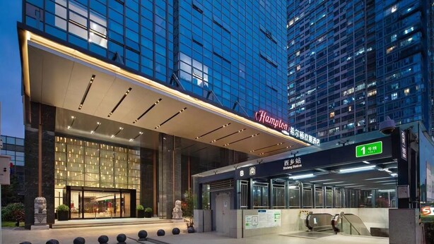 A modern hotel entrance with a glass facade, a prominent 'Hampton' sign, and an escalator leading to the entrance.