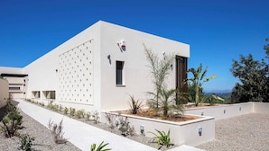 Un edificio moderno con techo plano, una ventana pequeña y una puerta. Hay plantas y un sendero de grava en frente.