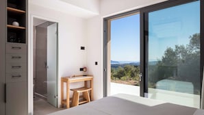 Un dormitorio moderno con un gran ventanal corredera que da a un balcón, un escritorio de madera y un armario con cajones.