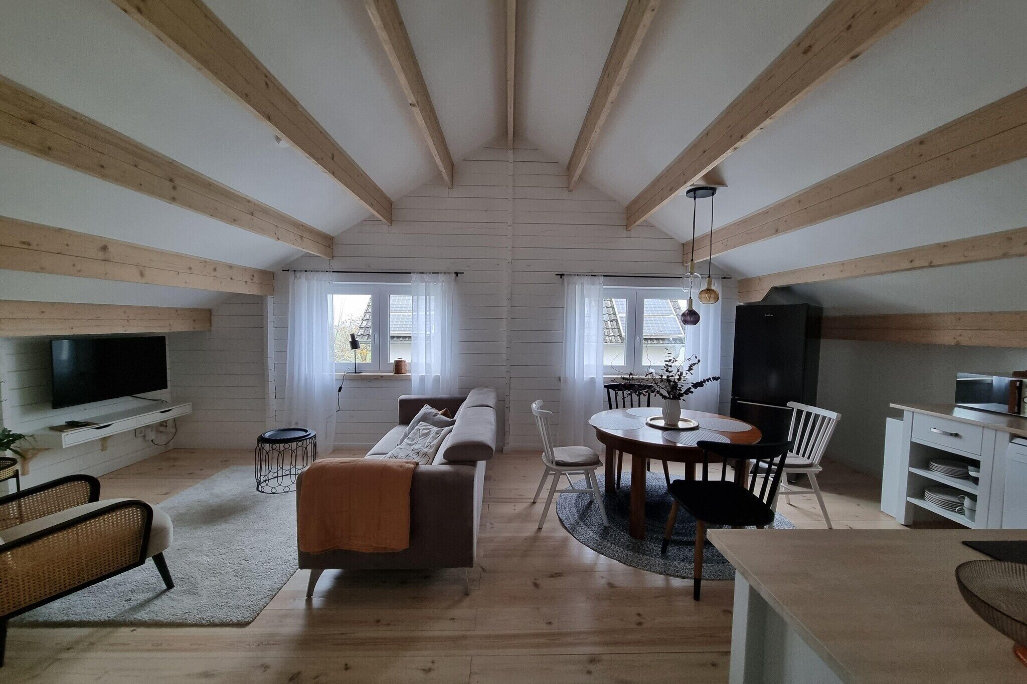 A spacious loft with exposed wooden beams, a large TV, a sofa, a dining table with chairs, and a kitchen area.