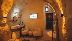 A cozy, vaulted room with a stone wall, a small desk with a chair, a television, and a radiator. - Daphne Cave Cappadocia (Avanos)