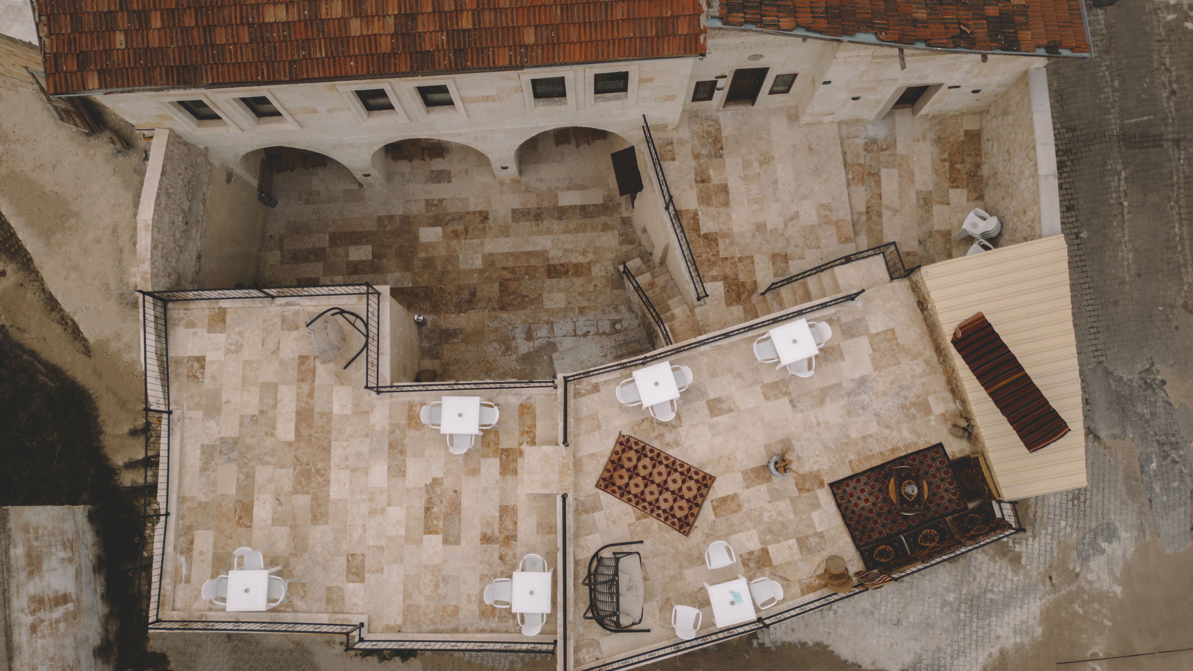 An aerial view of a courtyard with a tiled floor, white tables and chairs, a patterned rug, and a wooden bench.