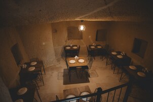 Un comedor con mesas dispuestas para cuatro, cada una con platos y cubiertos. La habitación tiene un aspecto rústico y similar a una cueva, con paredes de piedra y suelo de madera.