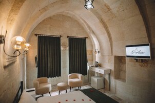 Una habitación de hotel con techo de piedra abovedado, una cama con alfombra verde, dos sillones, un escritorio con televisor de pantalla plana y un mueble pequeño.