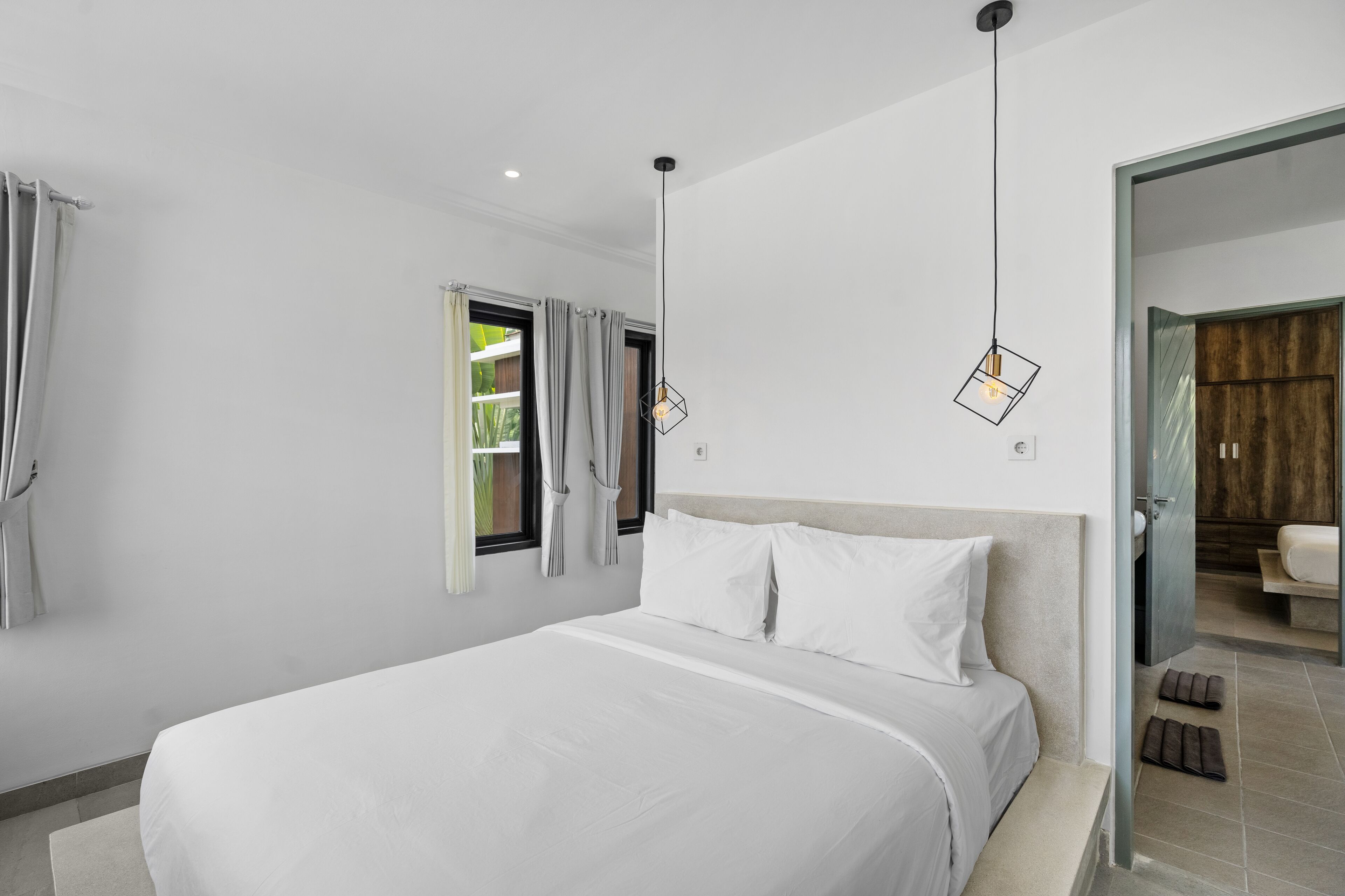 A modern hotel room with a large bed, a window with curtains, a hanging light fixture, and a view of the bathroom.