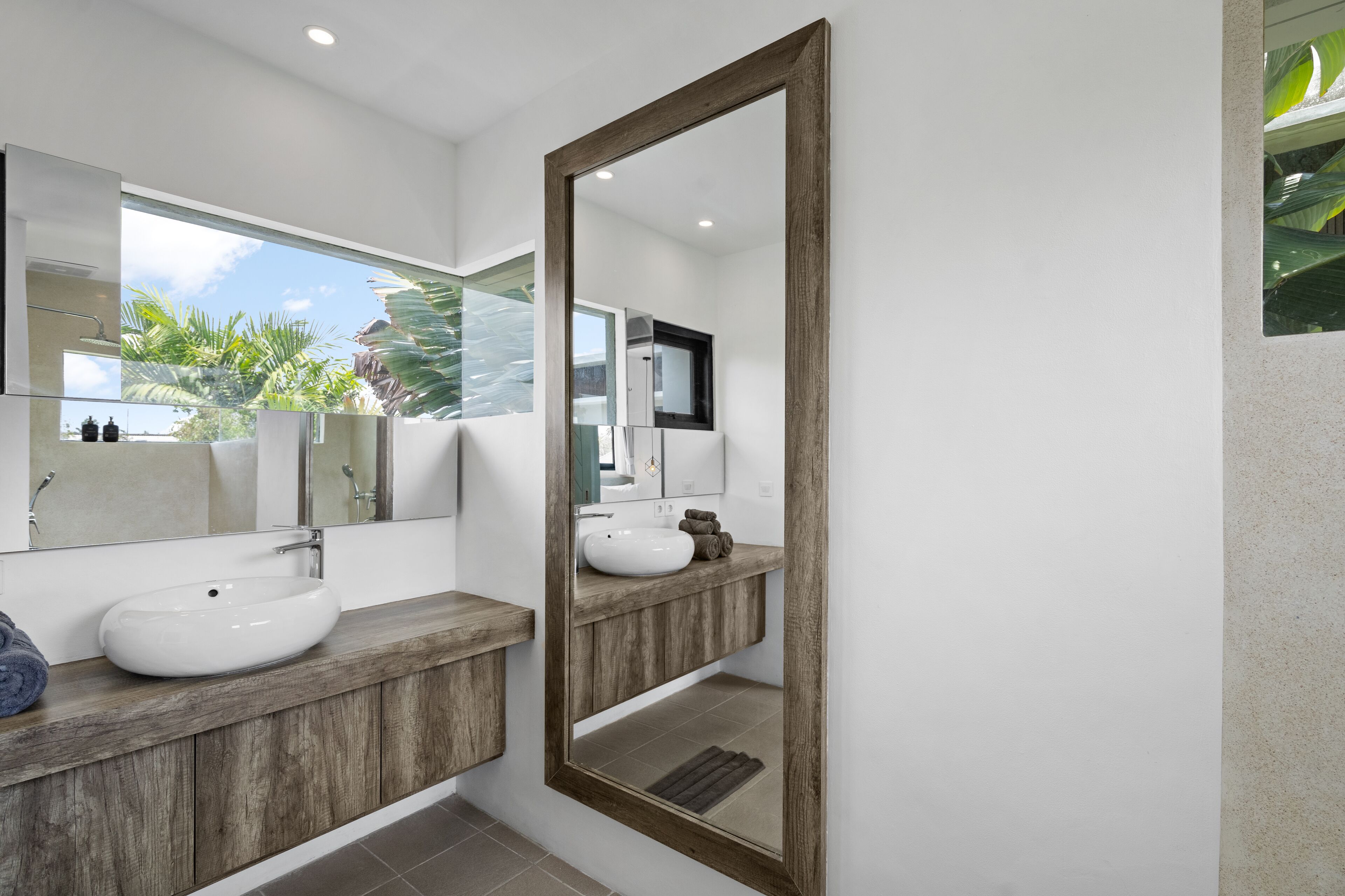 A modern bathroom with a large mirror, two sinks, wooden vanity, and a view of palm trees.