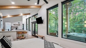 A modern bedroom with a large bed, a TV mounted on the wall, and a view of a hot tub through large windows. - Luxury Hocking Couples Cabin | Secluded! Hot Tub! (Logan)