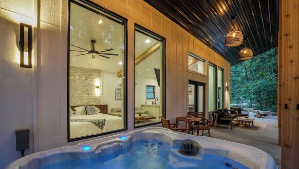 A modern spa area with a hot tub, wooden deck, and a bedroom with a large window. - Luxury Hocking Couples Cabin | Secluded! Hot Tub! (Logan)