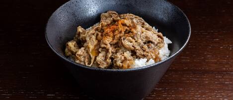A bowl of rice topped with a meat dish, possibly beef, in a dark bowl on a wooden surface.