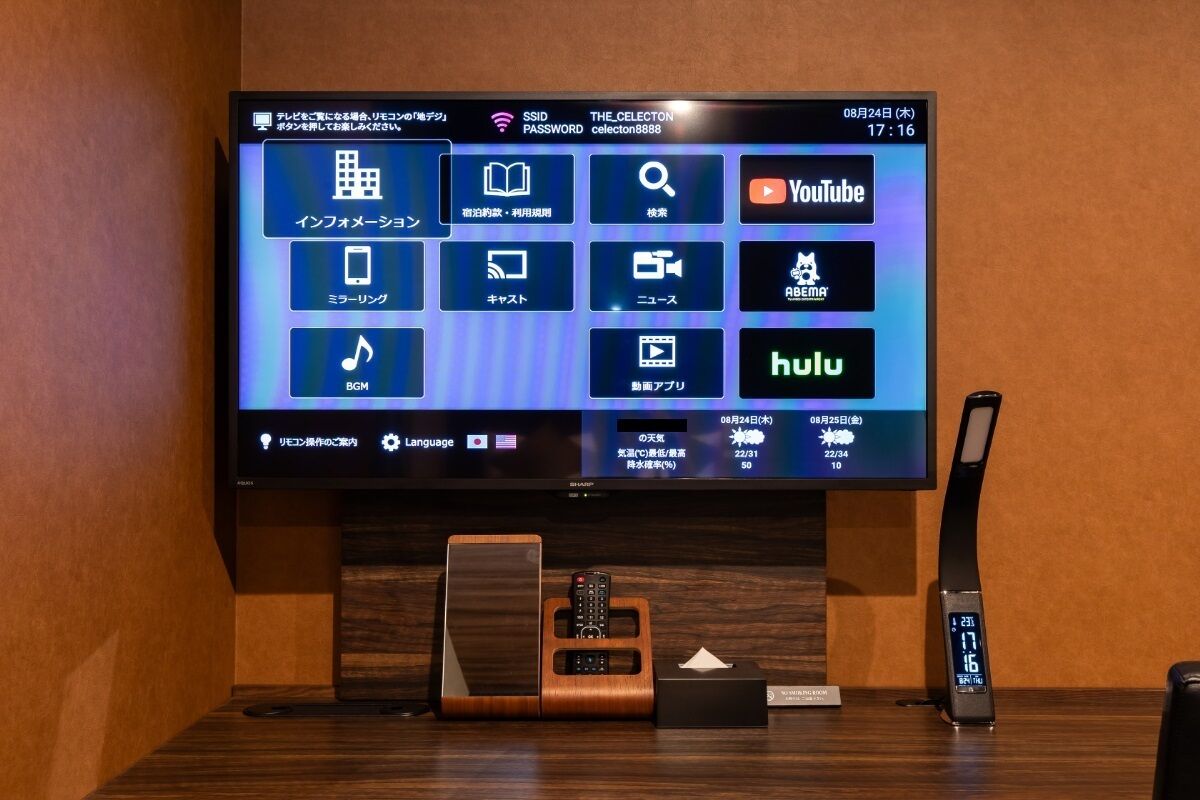 A hotel room with a flat-screen TV displaying various streaming services, a wooden side table with a remote control, a digital clock, and a small box.