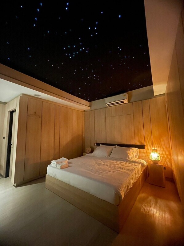 Un dormitorio con techo que simula un cielo estrellado, paredes de madera, una cama con sábanas blancas y una lámpara de noche.