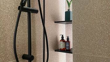 A shower with a handheld showerhead, a wall-mounted control panel, and a towel rack. There are two shelves with toiletries and a potted plant.