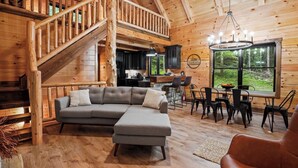 A wooden cabin interior with a staircase, a gray sectional sofa, a dining area, and a large window offering a view of the forest.