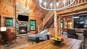 A cozy cabin interior with a stone fireplace, wooden walls, a staircase, and a dining area with a fruit bowl.